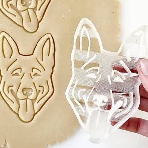 May include: A white plastic cookie cutter in the shape of a German Shepherd dog's head. The cutter is being held by a hand and is being used to cut out a cookie from a sheet of cookie dough.