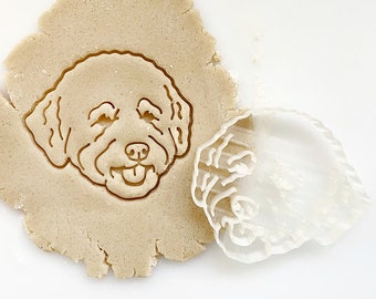 May include: A clear plastic cookie cutter in the shape of a dog's head is placed on top of a round piece of cookie dough. The cookie cutter is slightly offset from the center of the dough.