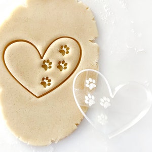 May include: A clear plastic heart-shaped cookie cutter with paw prints inside. The cutter is on top of a sheet of cookie dough with paw prints pressed into it.