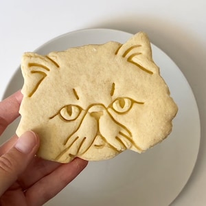 以下が含まれることがあります： 猫の顔の形をした薄い茶色の砂糖クッキー。クッキーには、猫の目、鼻、ひげを縁取る詳細な線が描かれています。