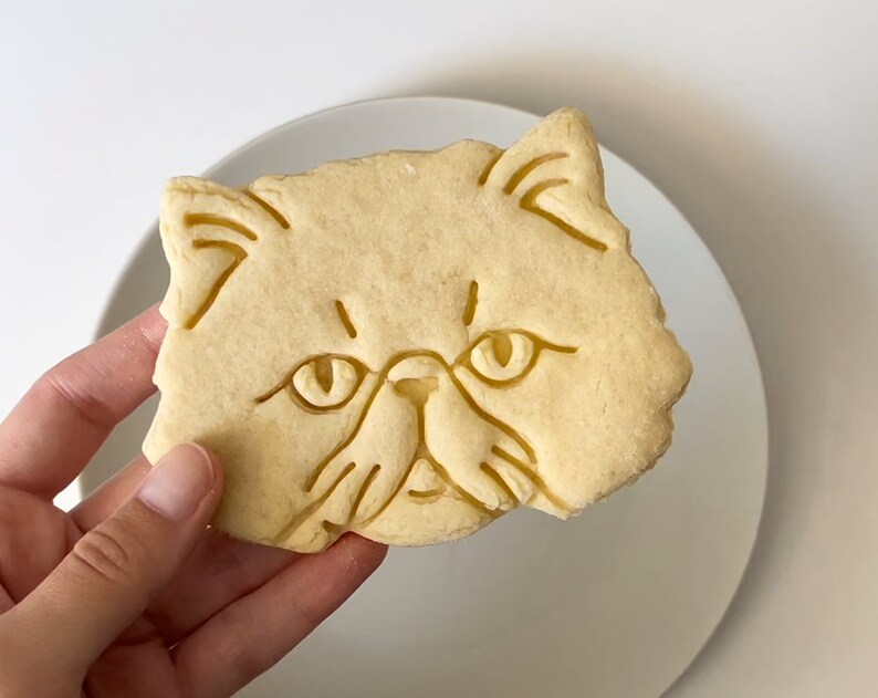 以下が含まれることがあります： 猫の顔の形をした薄い茶色の砂糖クッキー。クッキーには、猫の目、鼻、ひげを縁取る詳細な線が描かれています。