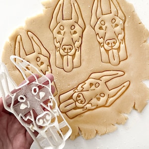May include: A white plastic cookie cutter shaped like a dog's head is held near pressed dough with dog head imprints. The dough is light brown, and the dog head imprints have detailed features.