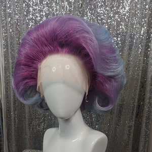 May include: A purple and blue wig with a lace front. The wig is styled in a bouffant with curls around the sides.