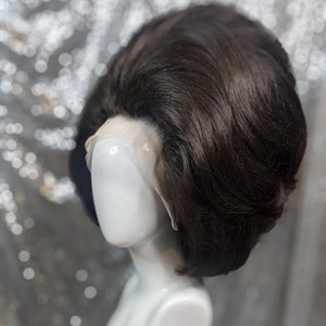 Może przedstawiać: Czarna peruka w stylu bouffant na białej głowie manekina. Peruka jest ułożona w krótkim, zaokrąglonym kształcie i ma gładkie, eleganckie wykończenie.