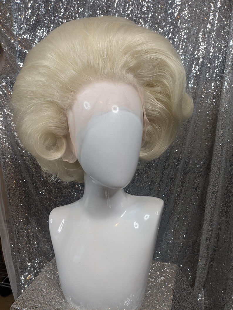 May include: A blonde wig with a bouffant style, styled with a side part and curled ends. The wig is on a white mannequin head.