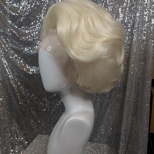 May include: A blonde wig with a side part and a voluminous, wavy style. The wig is on a white mannequin head.