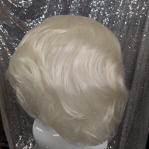 May include: A short, wavy, platinum blonde wig on a white mannequin head. The wig has a layered, choppy style.
