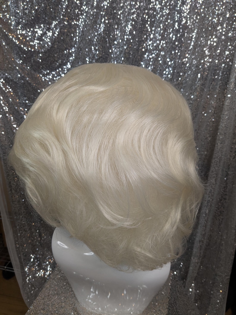 May include: A short, wavy, platinum blonde wig on a white mannequin head. The wig has a layered, choppy style.