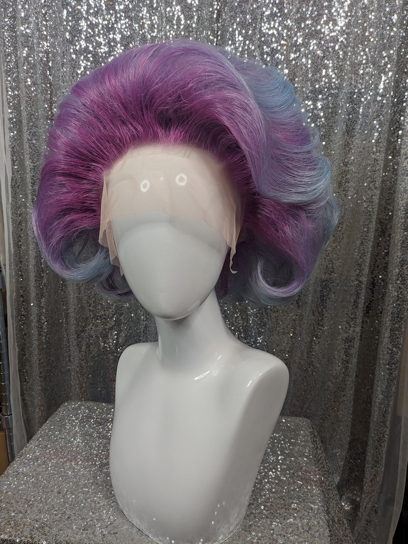 May include: A purple and blue wig with a lace front. The wig is styled in a bouffant with curls around the sides.