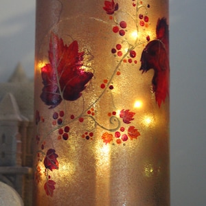 Könnte beinhalten: Ein goldener Glas-Kerzenhalter mit einem Rankendesign mit roten und orangefarbenen Blättern und Beeren. Der Kerzenhalter hat einen warmen Schein von den Lichtern im Inneren.