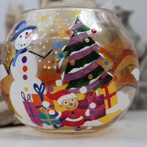 Może przedstawiać: Przezroczysta szklana kula z namalowaną zimową sceną. Kula przedstawia bałwana, choinkę i małą osobę trzymającą prezenty. Scena jest namalowana w żywych kolorach, w tym czerwonym, zielonym, niebieskim, żółtym i białym.