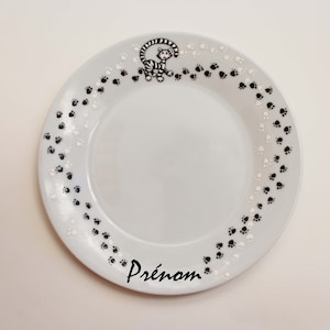 Könnte beinhalten: Weißer Keramikteller mit einem schwarzen und weißen Pfotenabdruckmuster am Rand. Oben auf dem Teller befindet sich eine schwarz-weiße Illustration einer Katze. Das Wort "Prénom" ist in Schwarz am unteren Rand des Tellers geschrieben.