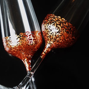 Könnte beinhalten: Zwei Champagnerflöten mit goldenen und kupfernen Malereien. Die Flöten befinden sich auf einem schwarzen Hintergrund.