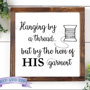 Könnte beinhalten: Ein weißes Schild mit einem braunen Holzrahmen, das eine schwarz-weiße Grafik einer Garnrolle und den Text "Hanging by a thread... but by the hem of HIS garment" zeigt.