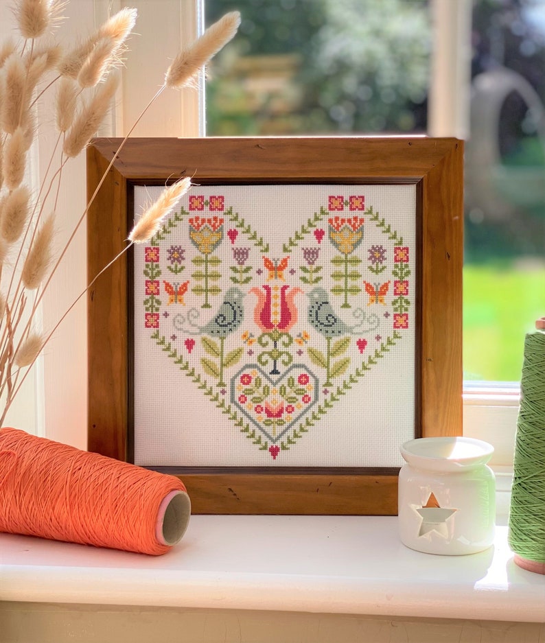 Puede incluir: Un dise&ntilde;o de punto de cruz enmarcado que presenta un patr&oacute;n en forma de coraz&oacute;n con dos p&aacute;jaros, flores y mariposas en tonos de verde, rosa, rojo, amarillo y azul. El marco est&aacute; hecho de madera con un acabado marr&oacute;n.