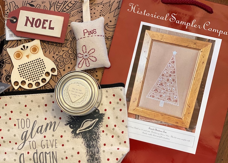 May include: A red bag with the text "Historical Sampler Company" and a framed cross-stitch design of a white Christmas tree with red and white ornaments. The bag also contains a variety of craft supplies, including a wooden owl, a small fabric pincushion with the word "Pins" embroidered on it, a small tin can with a heart-shaped design, and a fabric pouch with the text "Too glam to give a damn".