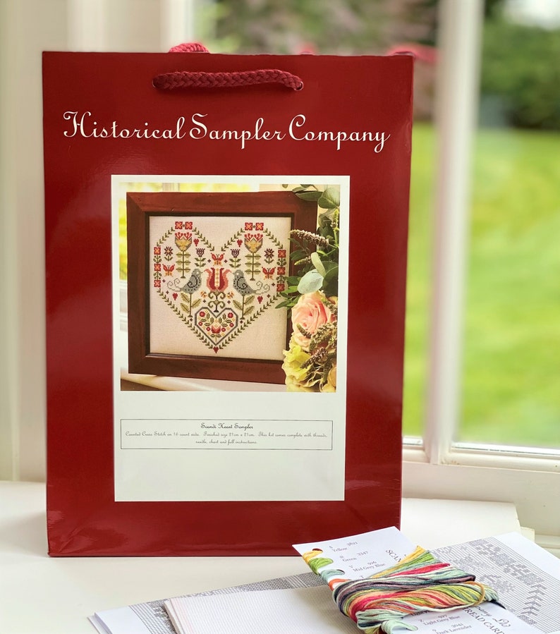 May include: A red gift bag with the text "Historical Sampler Company" printed on the front. The bag contains a cross stitch pattern with a heart design featuring two birds and flowers. The pattern is titled "Second Heart Sampler".