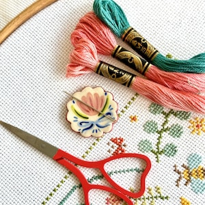 Può includere: Un tessuto bianco Aida con un motivo a punto croce floreale, un porta-ago in ceramica con un design floreale, un paio di forbici da ricamo rosse e tre bobine di filo da ricamo rosa e turchese.