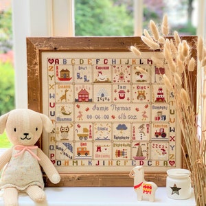 May include: A cross-stitch sampler in a wooden frame, featuring the alphabet and images like a giraffe, house, and tractor. The sampler includes the name "Jamie Thomas" and birth details. A soft toy and decorative items are placed in front of the sampler. The frame is rustic.