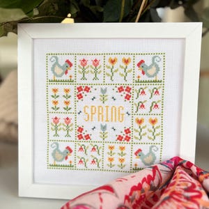 May include: A white framed cross-stitch embroidery with a spring theme. The design features a patchwork of colourful flowers, green leaves, and two blue chickens. The word "SPRING" is stitched in yellow in the centre of the design.