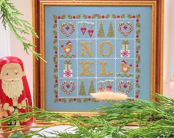 May include: A framed cross-stitch design with a blue background and a white border. The design features the word "NOEL" in yellow letters, surrounded by hearts, trees, and birds.