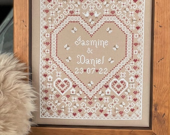 May include: A framed cross-stitch with a heart design and the names Jasmine & Daniel and the date 23 07 22. The heart is surrounded by a pattern of small hearts, birds, and flowers. The frame is a rustic brown wood.