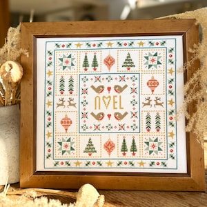 May include: A framed cross-stitch design with a white background and a variety of colourful Christmas motifs, including trees, stars, ornaments, and birds. The centre of the design features the word "NOEL" in gold lettering.