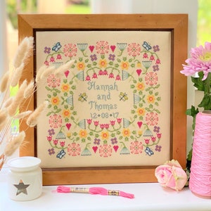 May include: A framed cross-stitch embroidery featuring a floral design and the text "Hannah and Thomas 12.08.17". The embroidery is in a wooden frame with a light brown finish. There is a pink embroidery floss, a white ceramic candle holder with a star design, and a pink rose on a white surface.