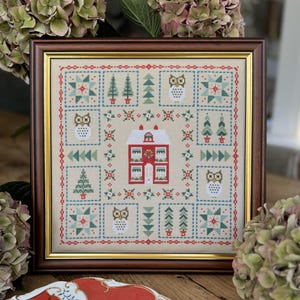 May include: A framed cross-stitch pattern featuring a red house, green trees, and white owls. The design is set against a cream background with a red, green, and white border.