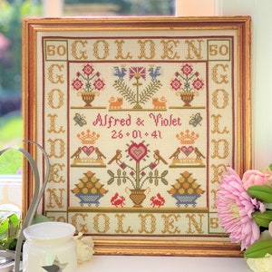 Könnte beinhalten: Ein gerahmter Kreuzstich-Sampler mit einem goldenen Rahmen und einem weißen Hintergrund. Der Sampler zeigt ein Blumenmuster mit Vögeln, Herzen und dem Text "50 GOLDEN 50" und "Alfred & Violet 26.01.41".