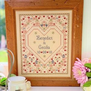 May include: A framed cross-stitch with a heart-shaped design featuring the names "Benedict & Cecilia" and the date "10-08-10". The cross-stitch is surrounded by a border of small hearts and flowers.