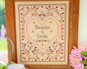 May include: A framed cross-stitch with a heart-shaped design featuring the names "Benedict & Cecilia" and the date "10-08-10". The cross-stitch is surrounded by a border of small hearts and flowers.