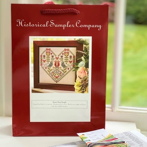 Puede incluir: Una bolsa de regalo roja con el texto "Historical Sampler Company" impreso en la parte delantera. La bolsa contiene un patr&oacute;n de punto de cruz con un dise&ntilde;o de coraz&oacute;n que presenta dos p&aacute;jaros y flores. El patr&oacute;n se titula "Second Heart Sampler".