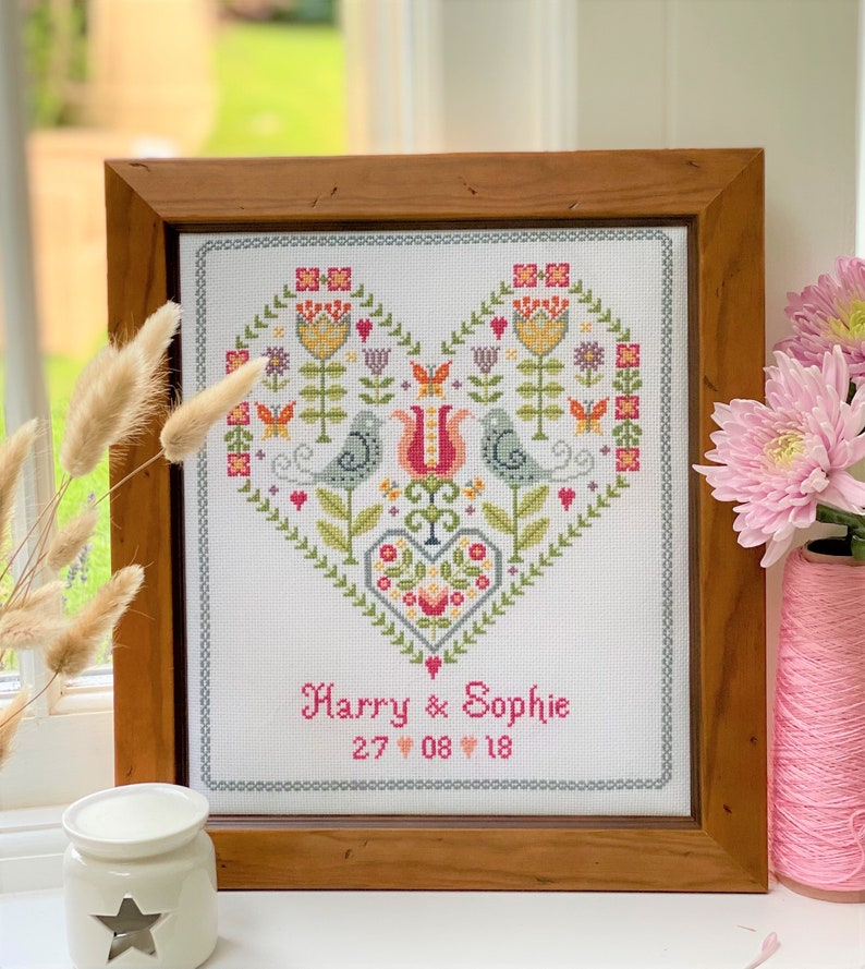May include: A framed cross-stitch design featuring a heart with stylized flowers, birds, and a smaller heart in the center. The text "Harry & Sophie 27 08 18" is below the heart.