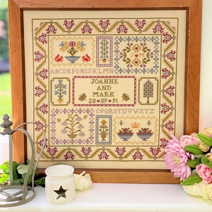 May include: A framed cross-stitch sampler with a cream background and colorful floral motifs. The sampler includes the names "Joanne and Mark" and the date "28.09.91". The alphabet is also included in the design.