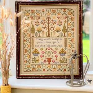 Peut inclure: Une broderie au point de croix encadrée avec un motif floral coloré et une citation : "Beaucoup d'eaux ne peuvent éteindre l'amour, ni les flots le noyer." La broderie est datée de 2001.
