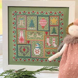 May include: A green cross-stitch sampler featuring a variety of Christmas-themed images, including a snowman, a Christmas tree, a stocking, and a star. The sampler is framed in white and is displayed on a table with a small doll.