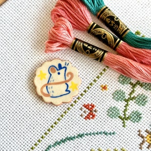 Könnte beinhalten: Eine Keramiknadel mit weißem Hintergrund und einer Comic-Maus mit blauem Schwanz und gelben Sternen. Die Nadel ist von einem braunen, gewellten Rand umgeben. Die Nadel liegt auf einem weißen Aida-Stoff mit grünem, blauem und rosa Stickgarn.