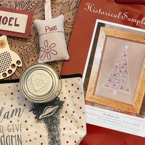 Puede incluir: Una bolsa roja con el texto "Historical Sampler Company" y un dise&ntilde;o de punto de cruz enmarcado de un &aacute;rbol de Navidad blanco con adornos rojos y blancos. La bolsa tambi&eacute;n contiene una variedad de art&iacute;culos de artesan&iacute;a, incluyendo un b&uacute;ho de madera, un peque&ntilde;o coj&iacute;n de tela con la palabra "Pins" bordada en &eacute;l, una peque&ntilde;a lata con un dise&ntilde;o en forma de coraz&oacute;n y una bolsa de tela con el texto "Too glam to give a damn".