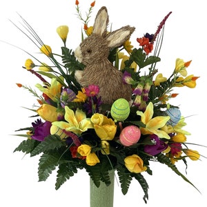 May include: A spring floral arrangement in a green vase features a brown bunny, yellow lilies, purple tulips, and colorful Easter eggs.