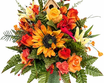 May include: A floral arrangement featuring a large central sunflower, orange and yellow roses, and green fern leaves. Two red cardinal bird figures are perched within the flowers, and a small wooden birdhouse is nestled at the top center.