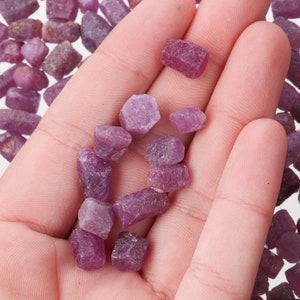 Könnte beinhalten: Eine Hand hält eine Sammlung kleiner, rauer, violetter Rubin-Edelsteine. Die Steine sind in verschiedenen Formen und Größen.