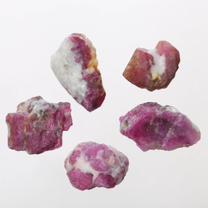 May include: Five rough, pink and white ruby gemstones. The gemstones are various shapes and sizes.