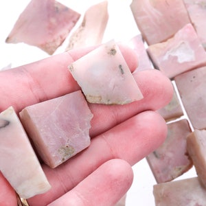 Könnte beinhalten: Eine Sammlung von rohen rosa Opal-Steinen, einige sind quadratisch, andere unregelmäßig. Die Steine sind in verschiedenen Größen und haben eine durchscheinende Optik.