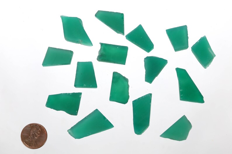 May include: A collection of 15 green gemstone chips, some with a rectangular shape and others with a triangular shape. The chips are scattered on a white surface, with a US penny for scale.