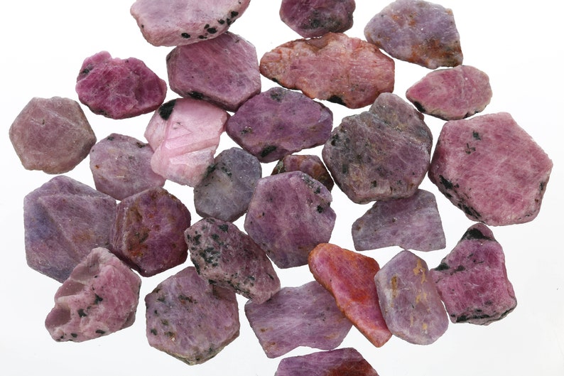 May include: A collection of pink and purple rough ruby gemstones. The stones are various sizes and shapes, with some having a more pointed or angular shape.