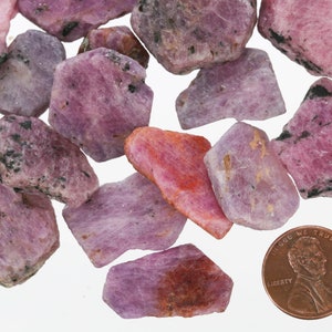 May include: A collection of pink and purple rough ruby gemstones, some with a darker, almost black, inclusion. The gemstones are scattered on a white surface, with a UK penny for scale.
