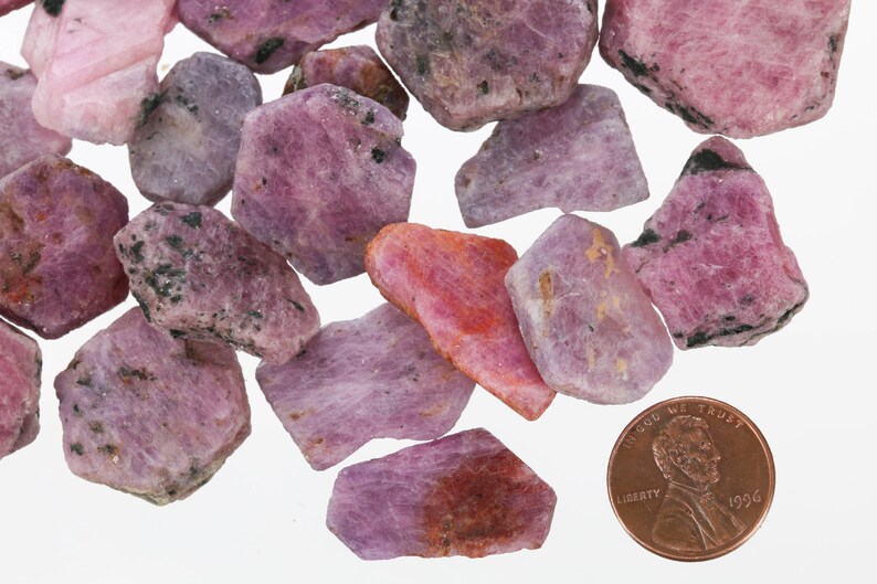 May include: A collection of pink and purple rough ruby gemstones, some with a darker, almost black, inclusion. The gemstones are scattered on a white surface, with a UK penny for scale.