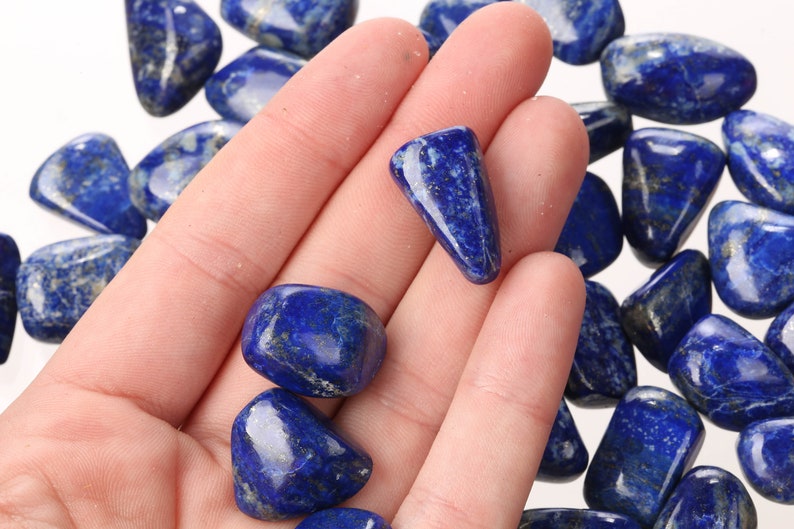 May include: A hand holding a collection of tumbled lapis lazuli stones. The stones are a deep blue color with flecks of gold.