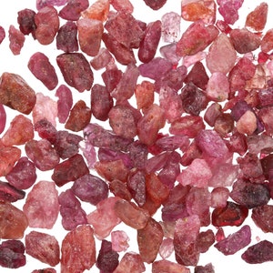 May include: A collection of small, rough, pink and red gemstones. The gemstones are scattered on a white background.